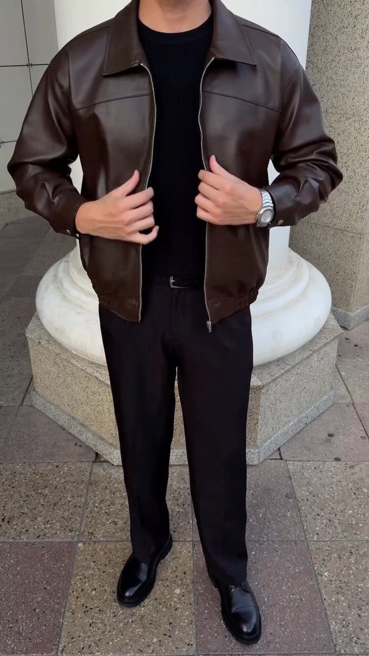 Man wearing a brown leather jacket, black t-shirt, black trousers, and black loafers standing outdoors.
