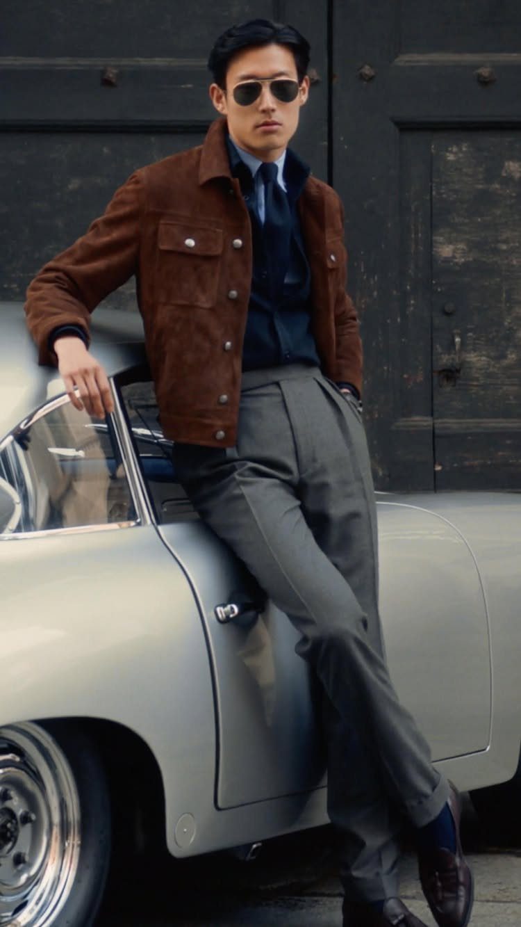 Man in a brown suede jacket and grey trousers leaning against a vintage silver car.