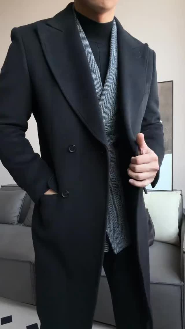 Man wearing a long dark overcoat, patterned waistcoat, and black turtleneck sweater indoors.