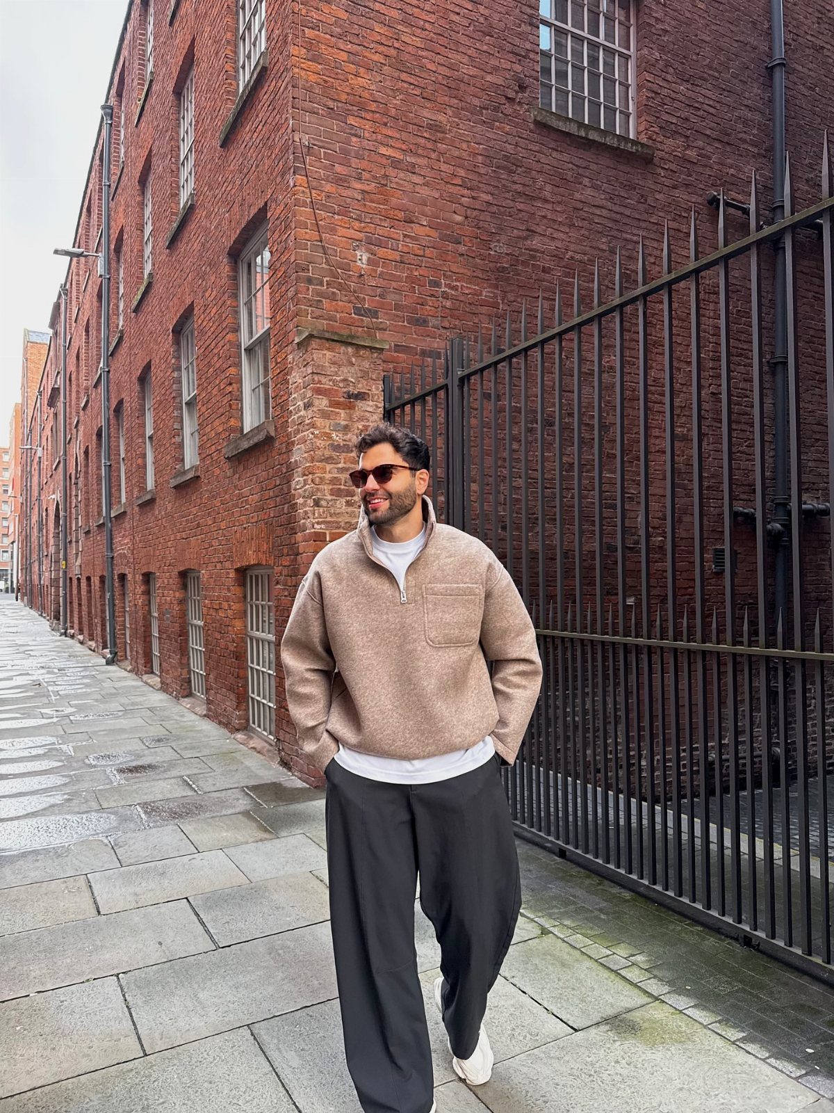 A man wearing a tan fleece and black wide-leg trousers walking on a city sidewalk.