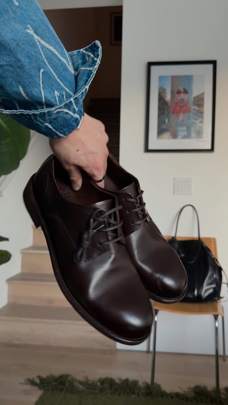 A hand holding up a pair of polished dark brown leather derby shoes indoors.