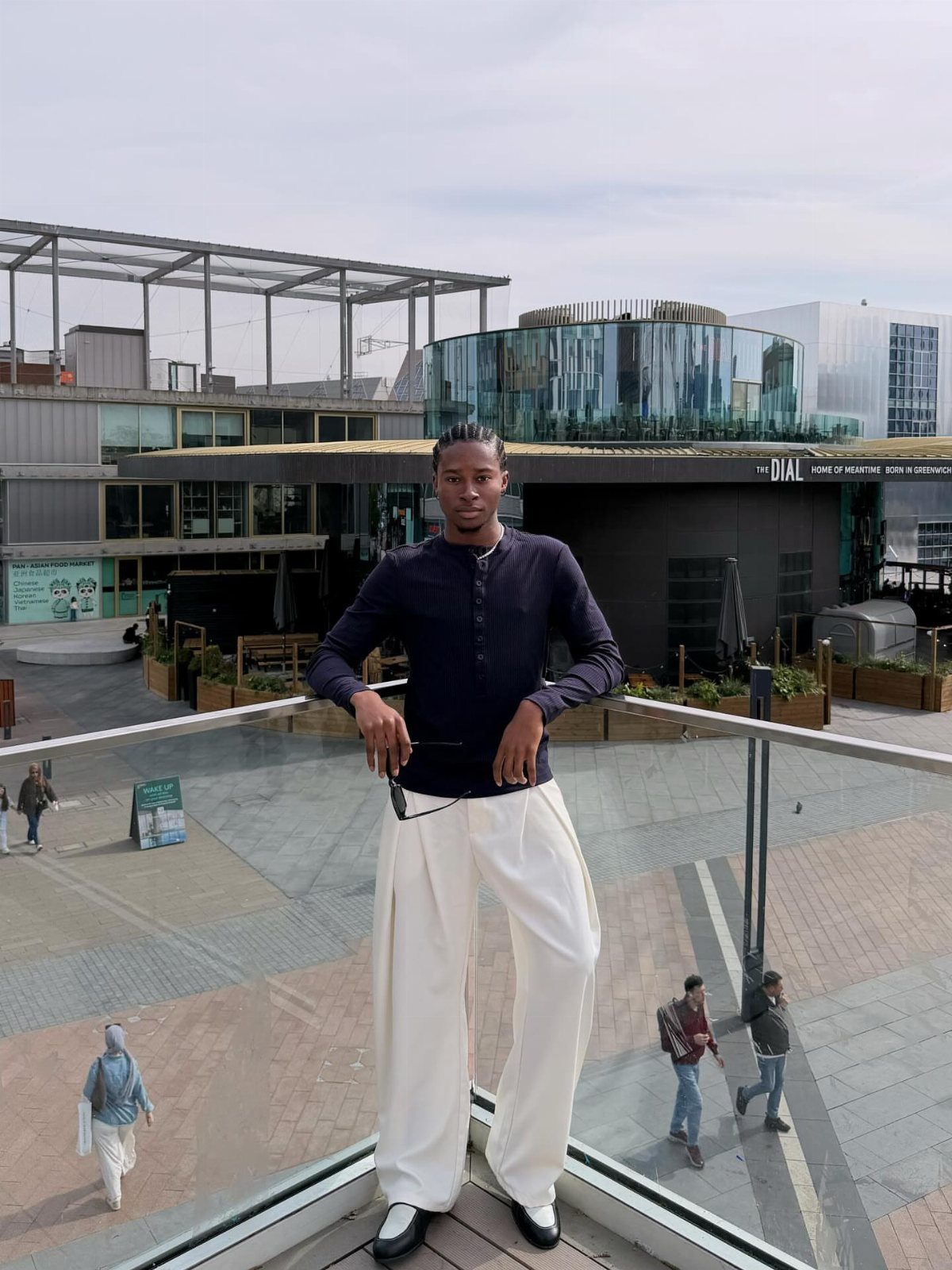 A man wearing a dark ribbed henley shirt and wide-leg white trousers stands on a glass balcony.
