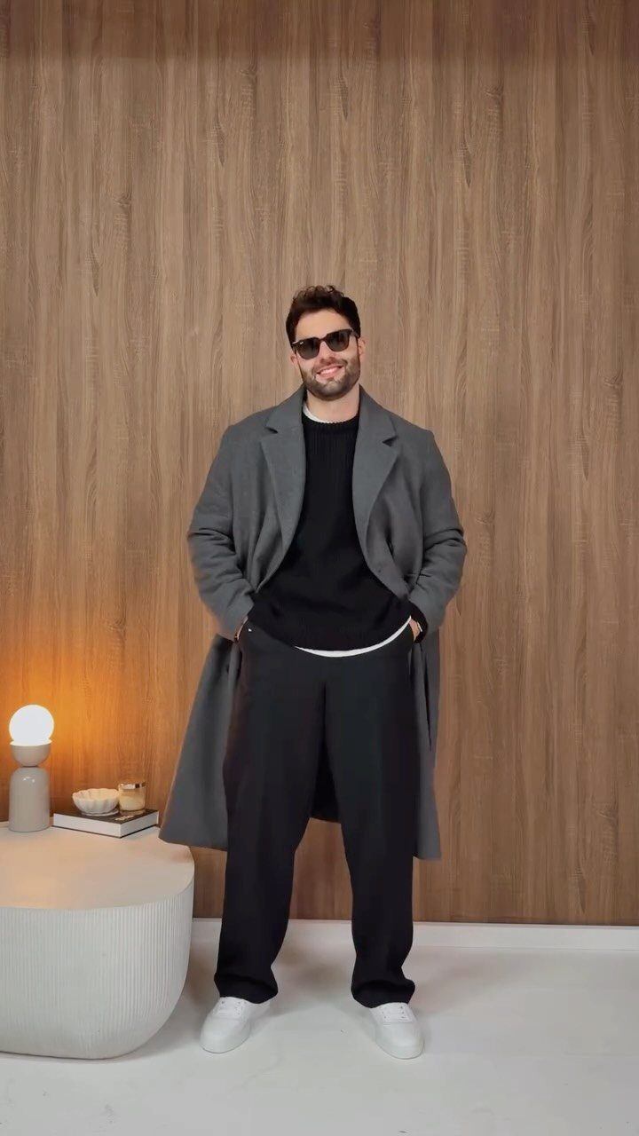 A smiling man in a grey coat, black sweater, and black trousers standing against a wood wall.