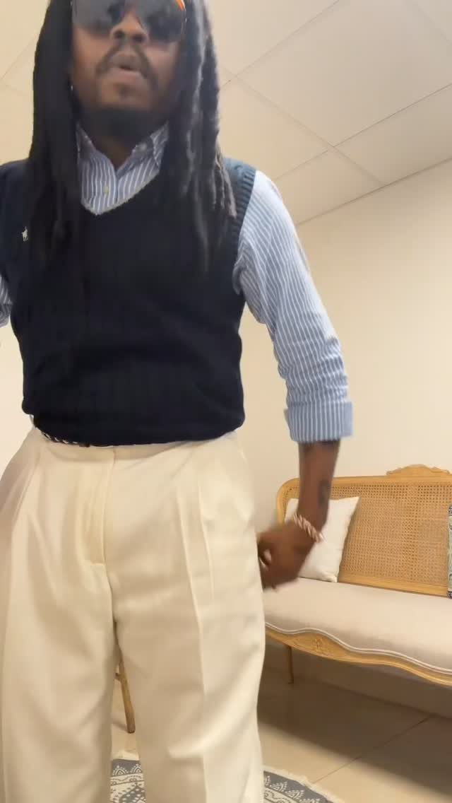 Man with dreadlocks wearing a sweater vest, striped shirt, and cream trousers standing indoors.