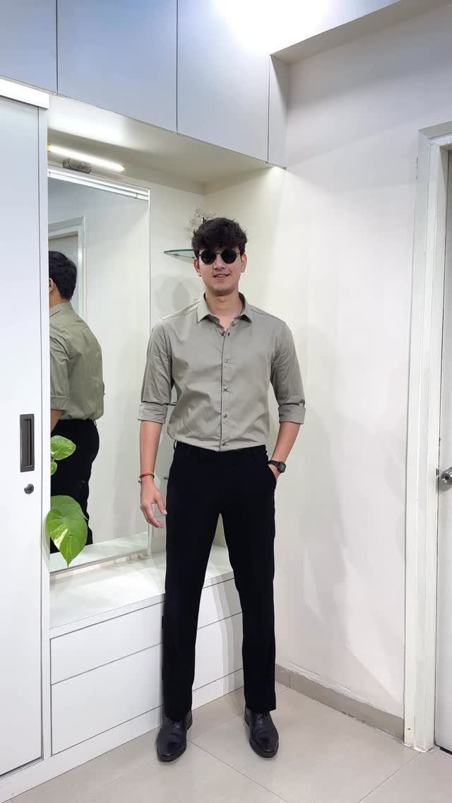 A man wearing a grey button-down shirt, black trousers, and sunglasses posing indoors.