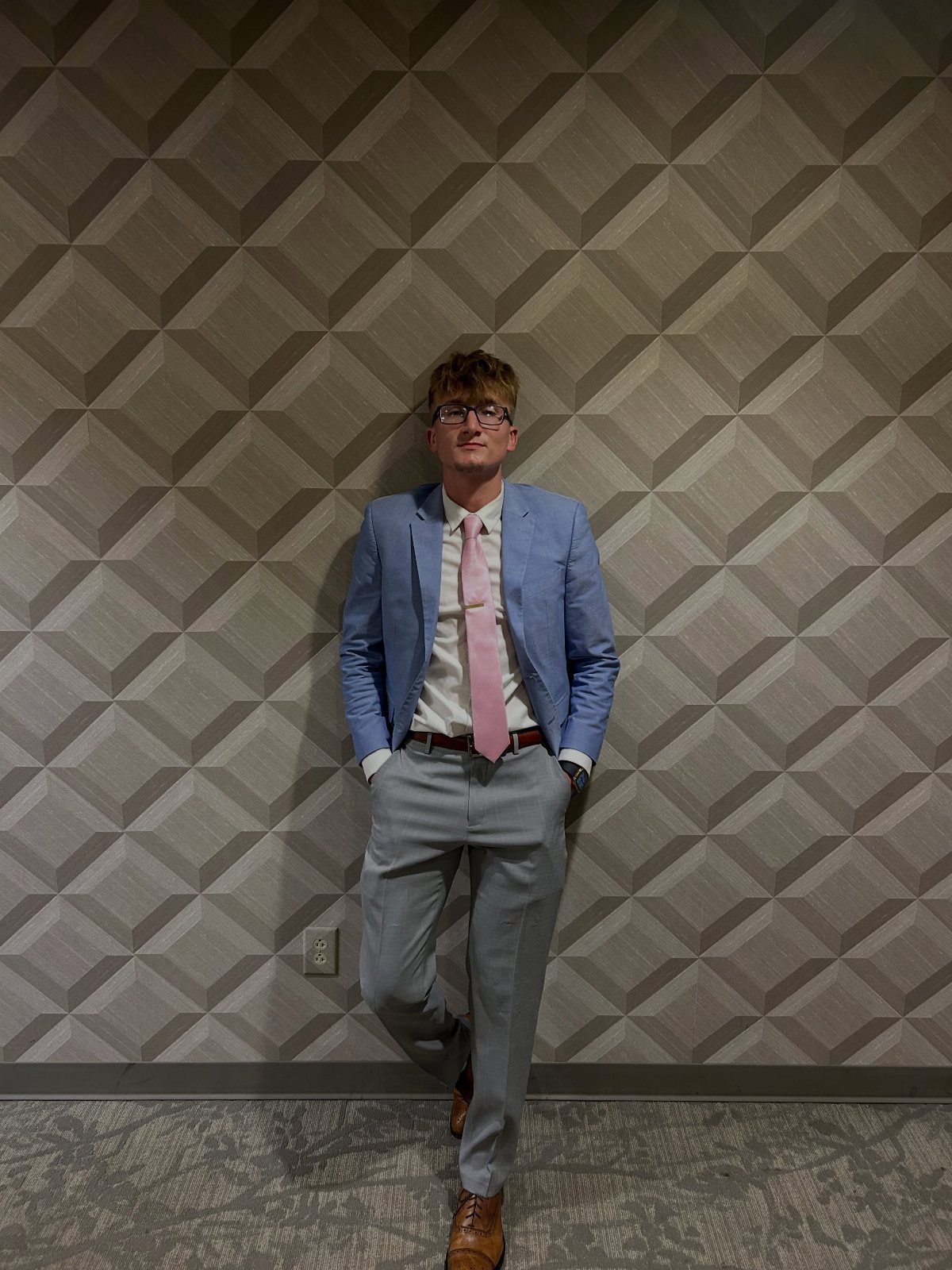 Man in a light blue blazer, pink tie, and grey trousers leaning against a patterned wall.