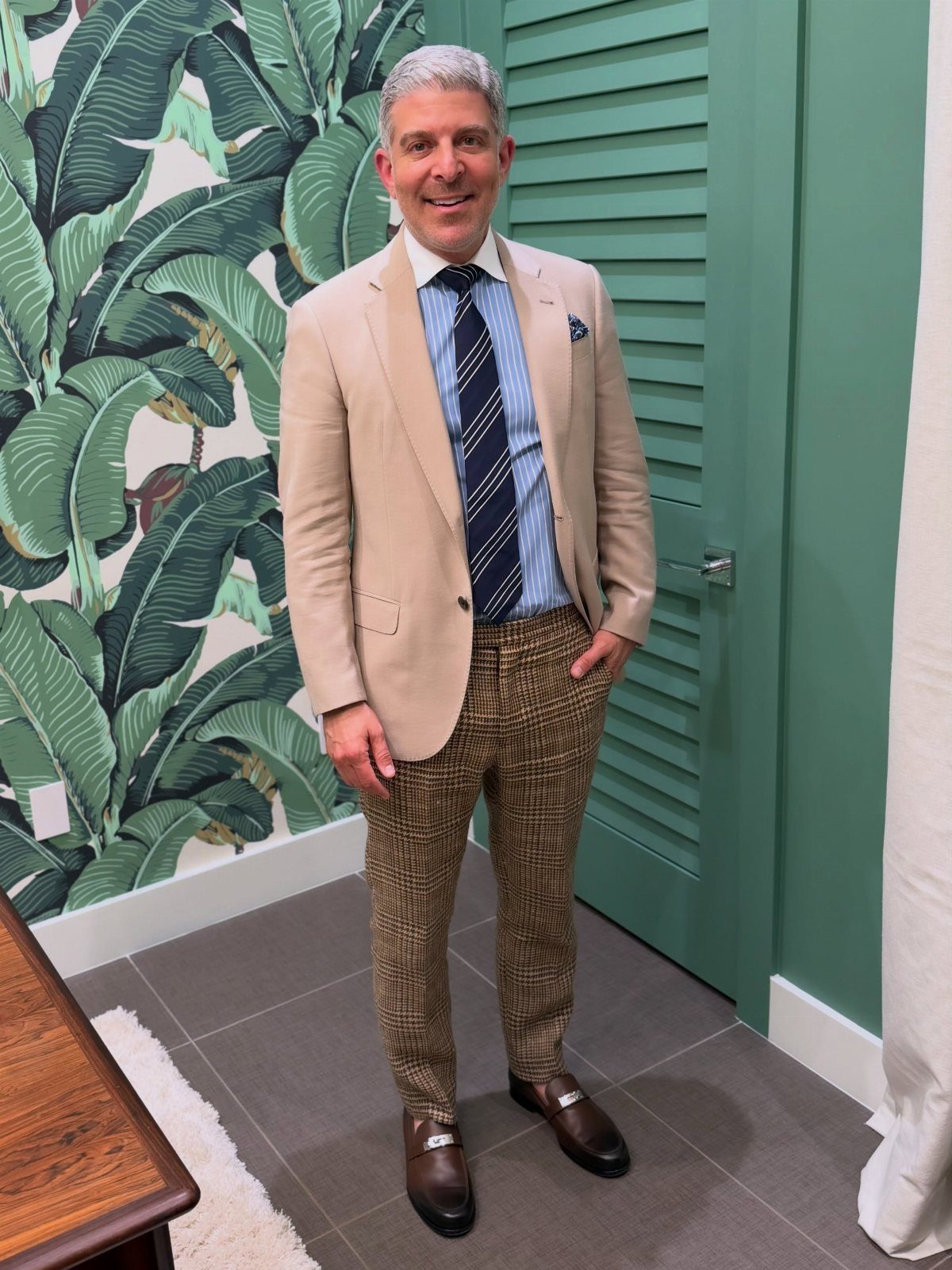 Man in a tan blazer, striped shirt, patterned tie, and brown trousers standing indoors.