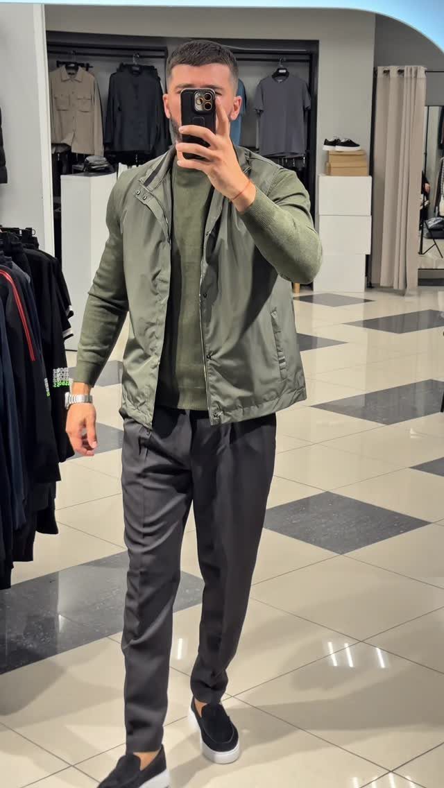 Man in olive green jacket and sweater with dark grey trousers standing in a clothing store.
