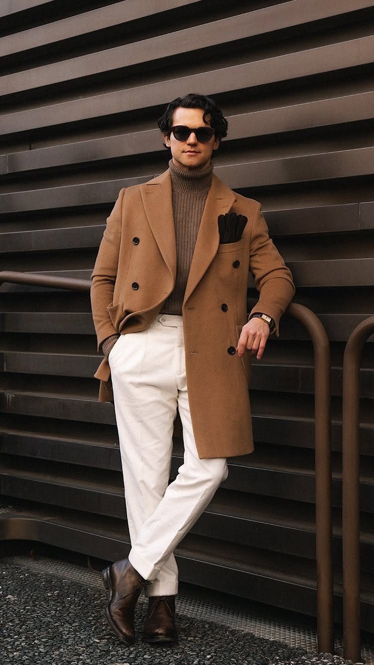 Man in tan overcoat, brown turtleneck, and white trousers posing against a dark slatted wall.
