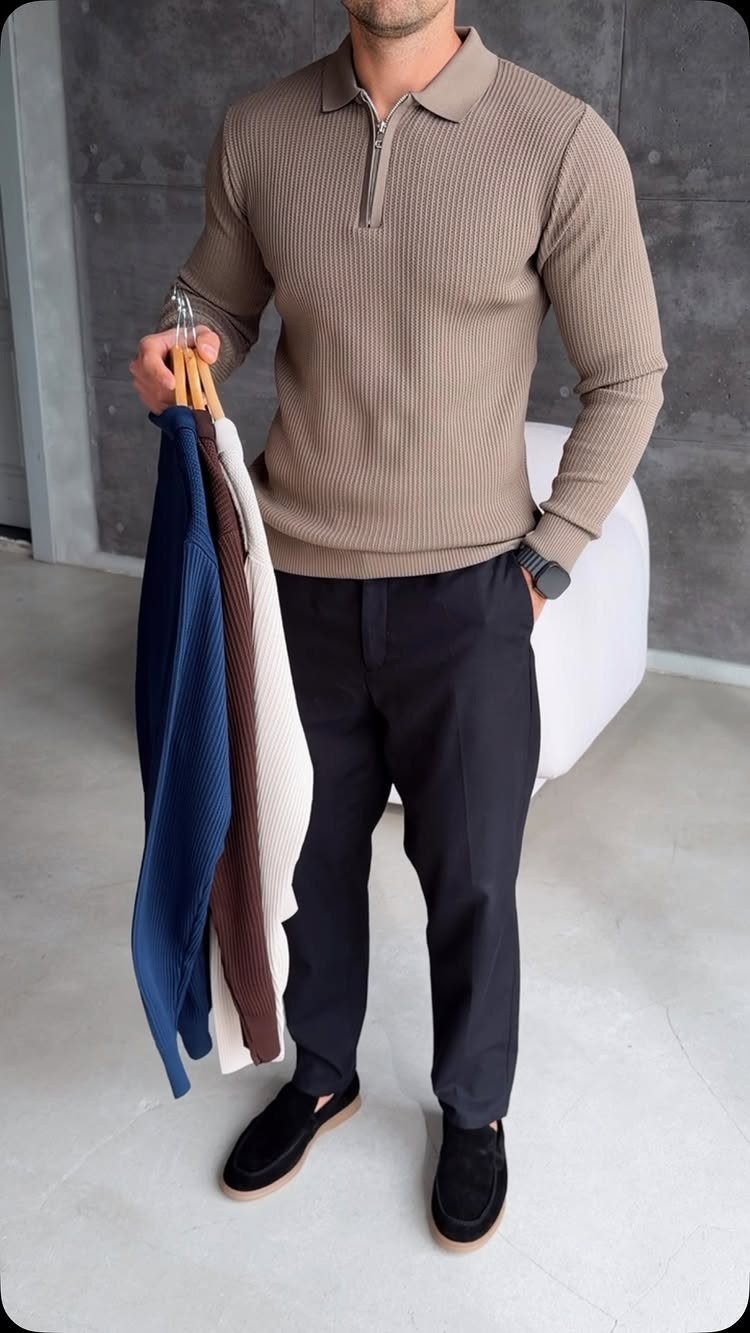 Man in a tan ribbed quarter-zip shirt and dark trousers holding various textured garments on hangers.