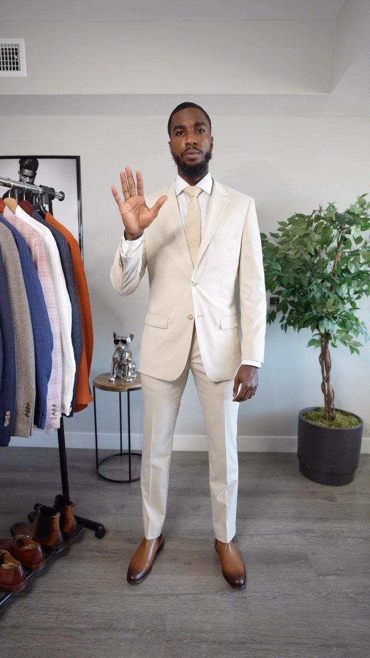 A man posing in a light beige tailored suit and gold tie in a bright room.