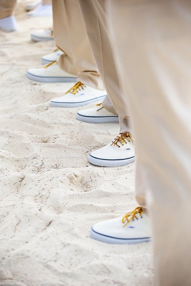 Several people wearing white canvas sneakers with yellow laces standing on a sandy beach.