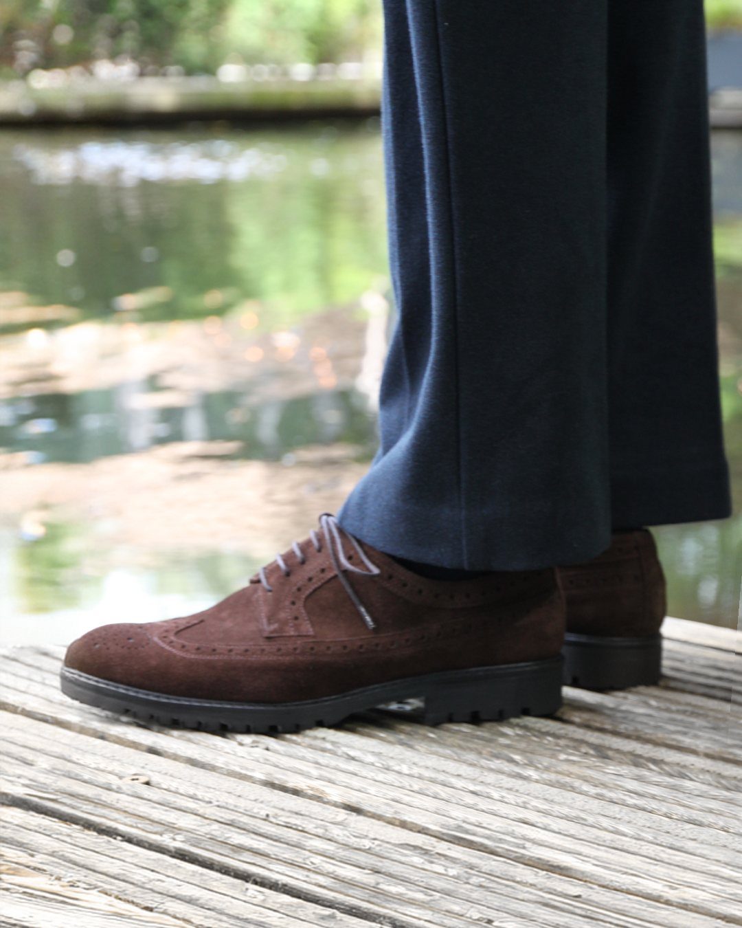 Brown suede brogue shoes worn with navy blue trousers on a wooden deck.