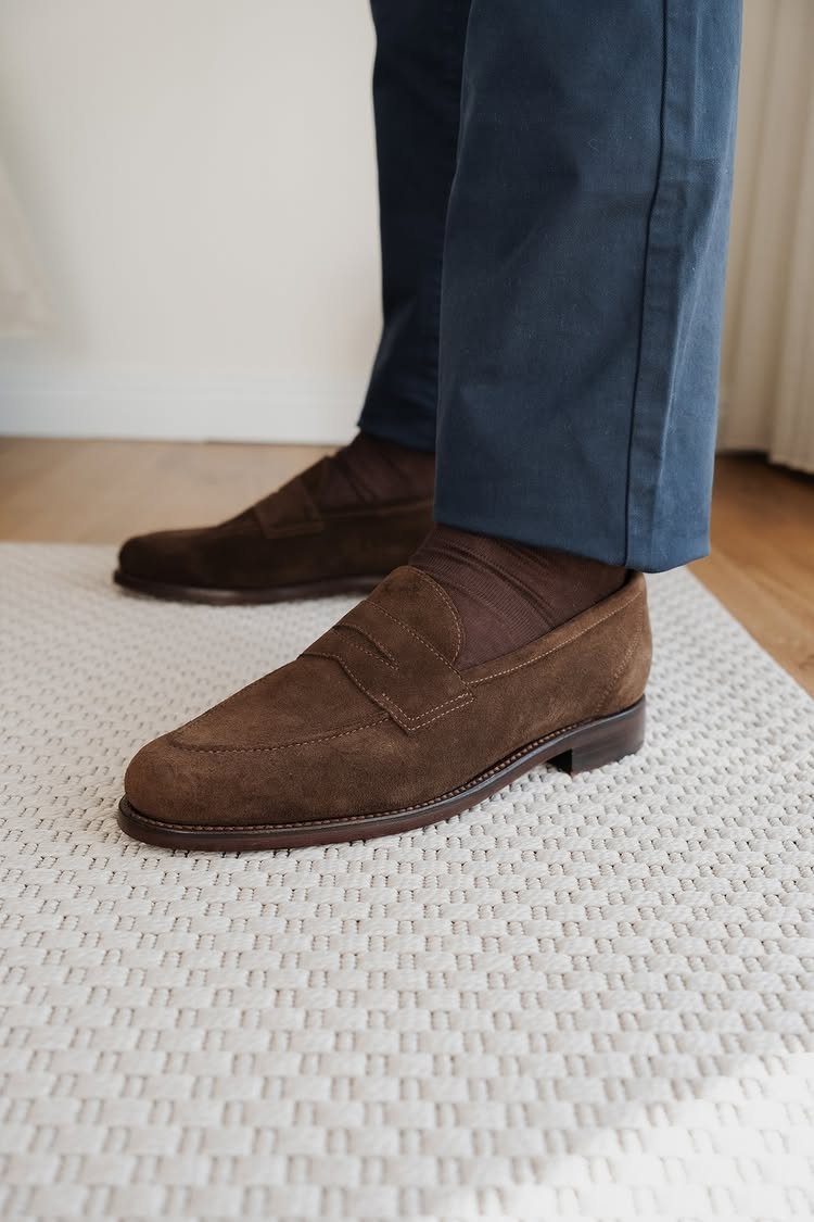 Brown suede loafers and blue trousers worn by a person standing on a white rug.