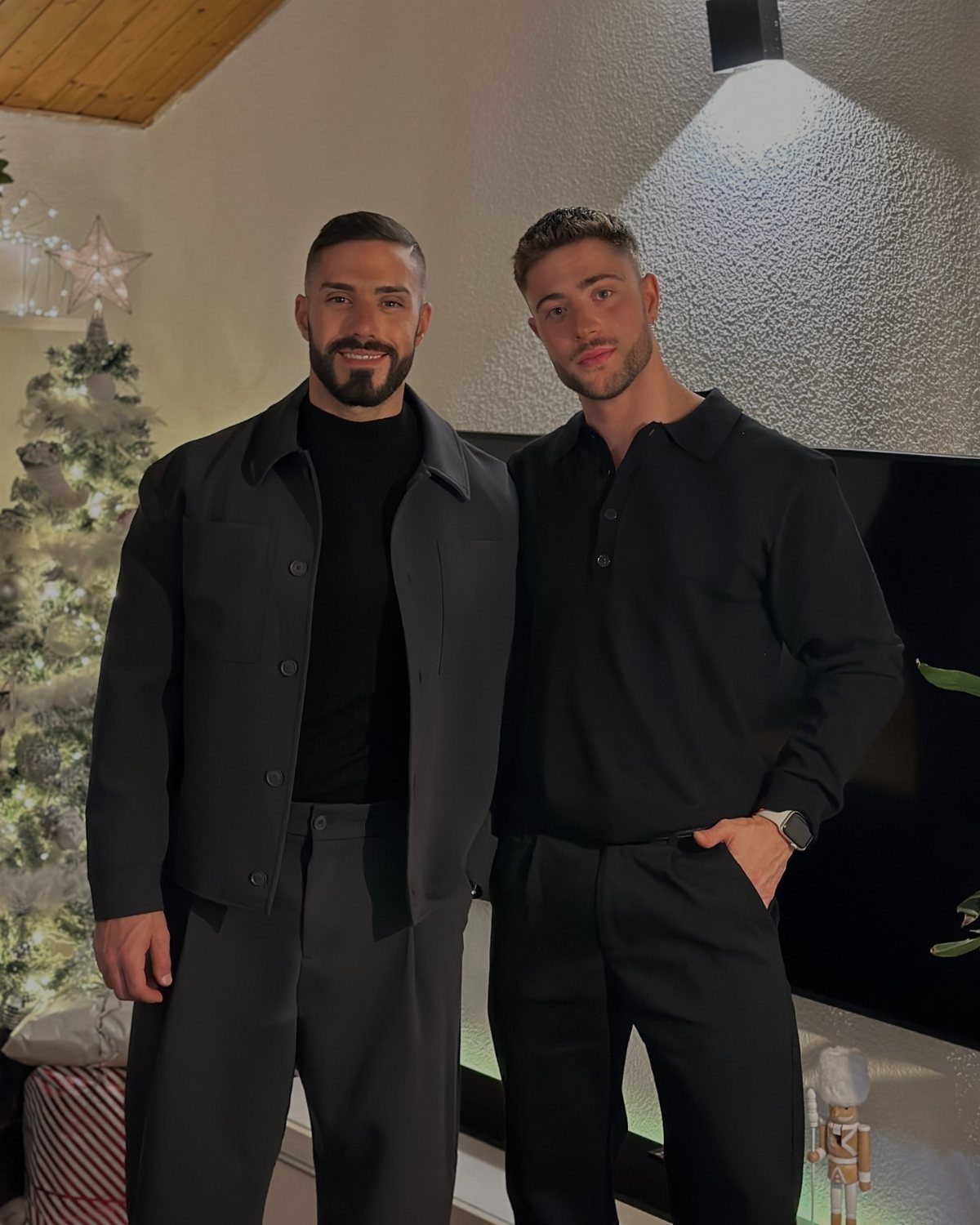 Two men posing in stylish dark clothing next to a decorated Christmas tree.