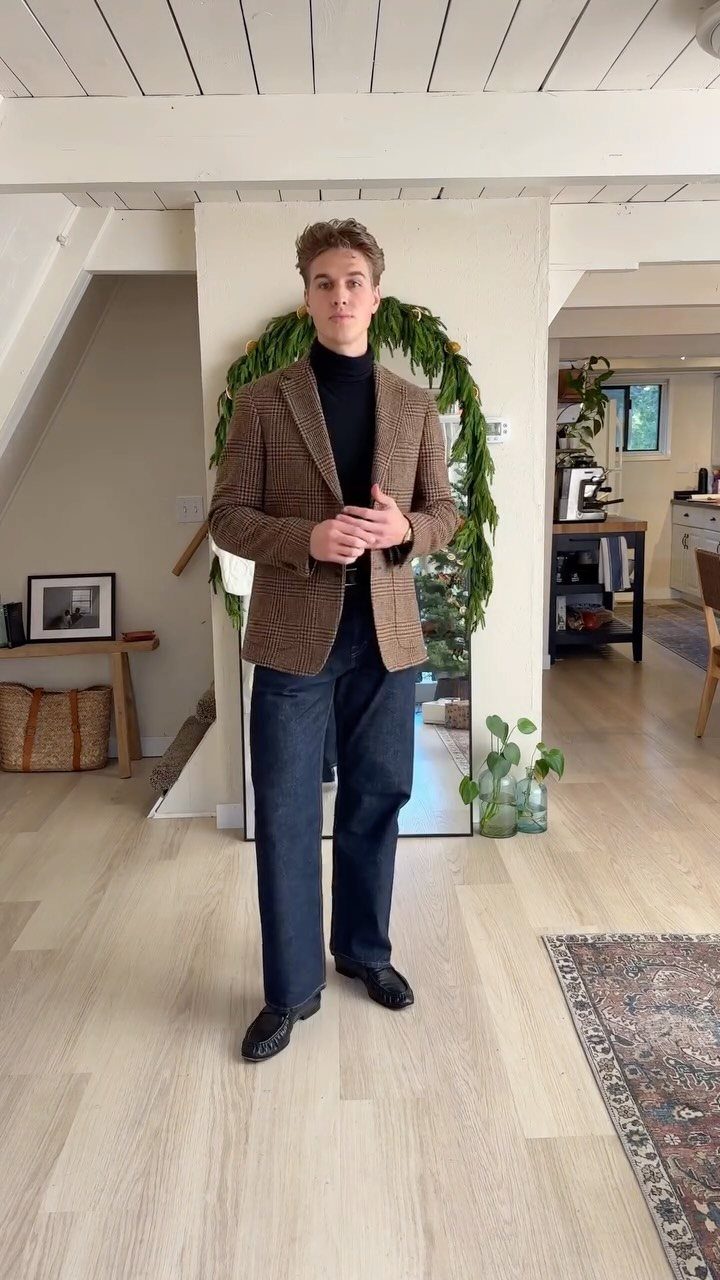 A young man posing in a brown plaid blazer, black turtleneck, and dark blue trousers.