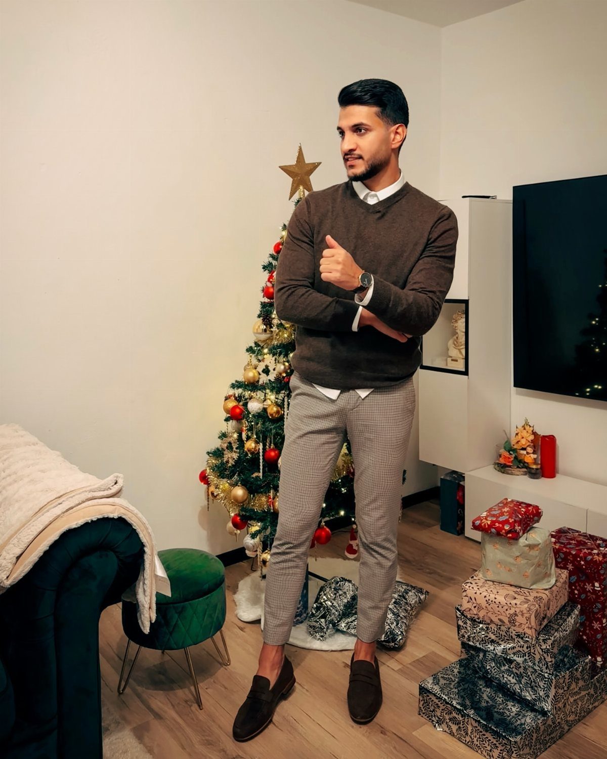 Man wearing a brown sweater and patterned trousers standing near a decorated Christmas tree.