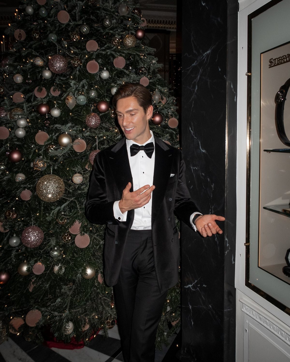 Man wearing a black velvet tuxedo and bow tie standing by a Christmas tree.