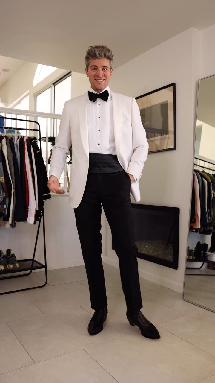 A man smiling while wearing a white tuxedo jacket with a black bow tie and trousers.