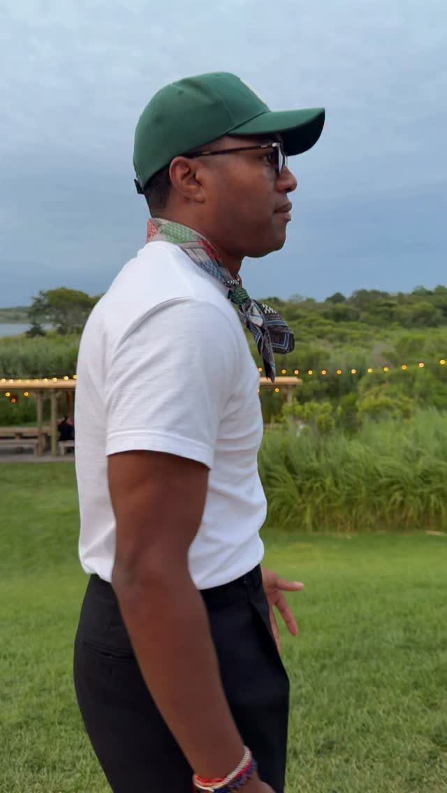 Man in a white t-shirt, green cap, and patterned scarf standing on a grassy lawn.