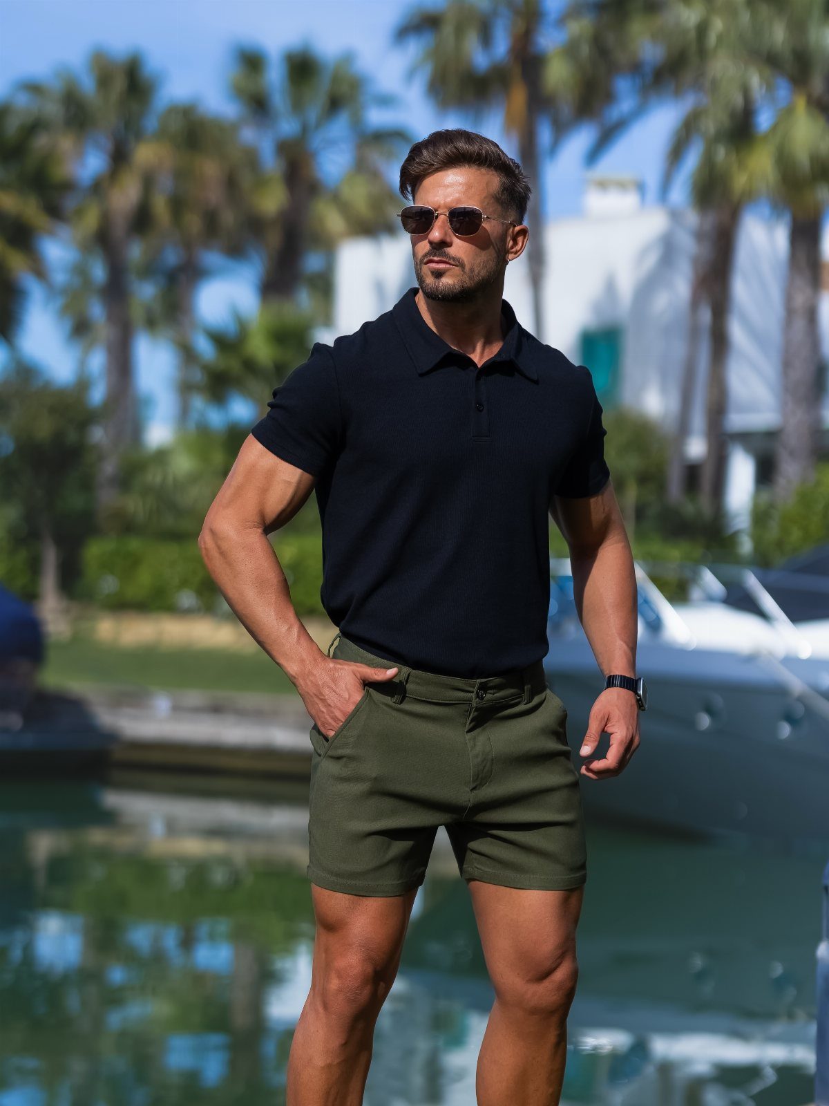 Man in black polo and olive green shorts standing by a tropical waterfront.
