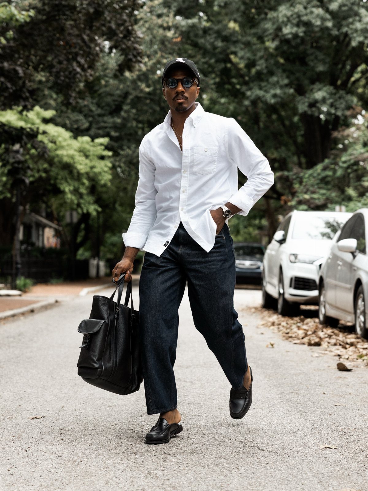 Man in white button-down shirt and dark denim walking with a black leather bag.