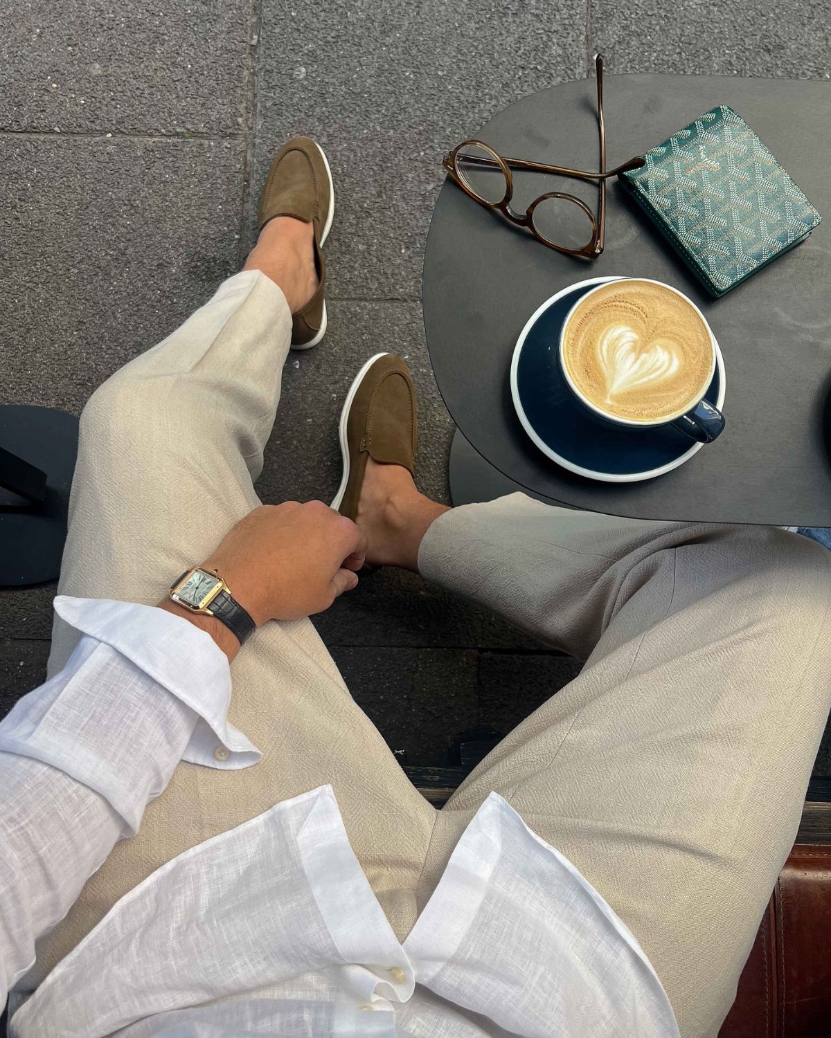 Person wearing beige linen trousers and brown suede loafers sitting at an outdoor cafe table.