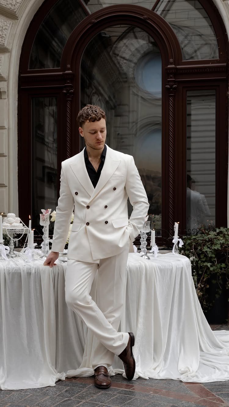 Man in a white double-breasted suit and black shirt leaning against a table outdoors.