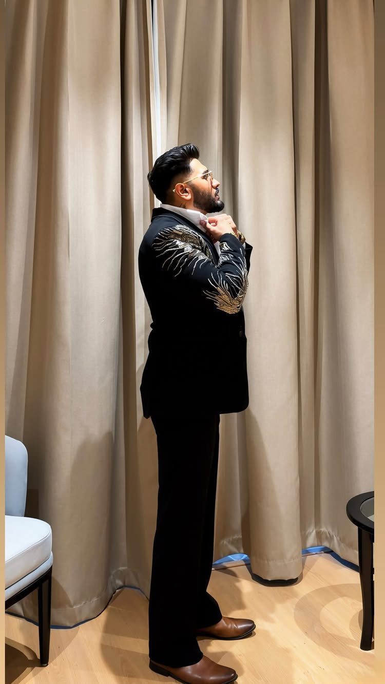 A man in a black patterned blazer and trousers standing against beige curtains.