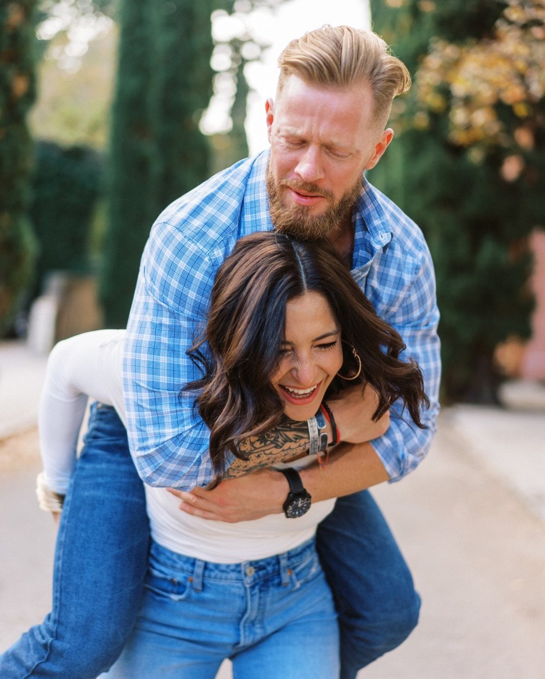 A man in a blue plaid shirt gives a smiling woman a piggyback ride outdoors.