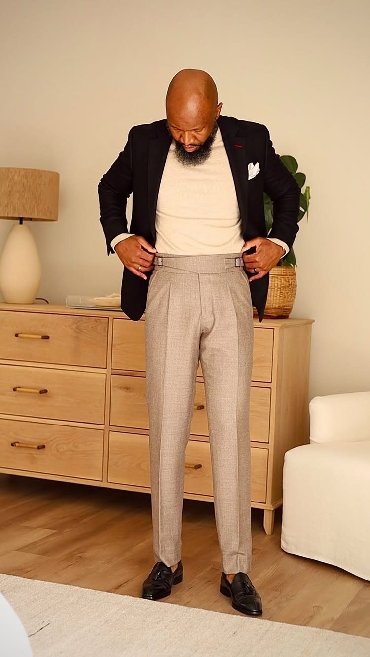 A man in a black blazer, cream sweater, and grey trousers stands in a room.