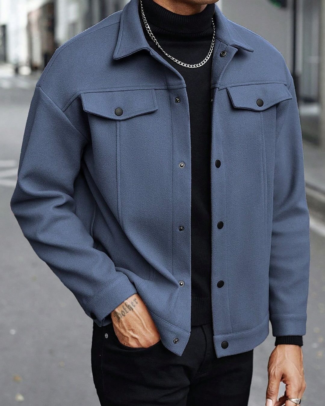 A man wearing a blue jacket, black turtleneck, and silver chain necklace outdoors.