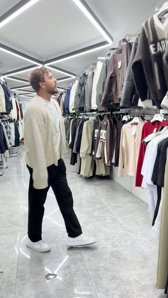 Man in a cream cardigan, white t-shirt, black pants, and white sneakers standing in a clothing store.