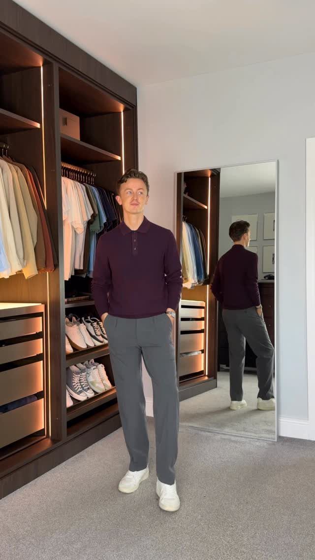 A man posing in a purple polo shirt and grey trousers inside a walk-in closet.