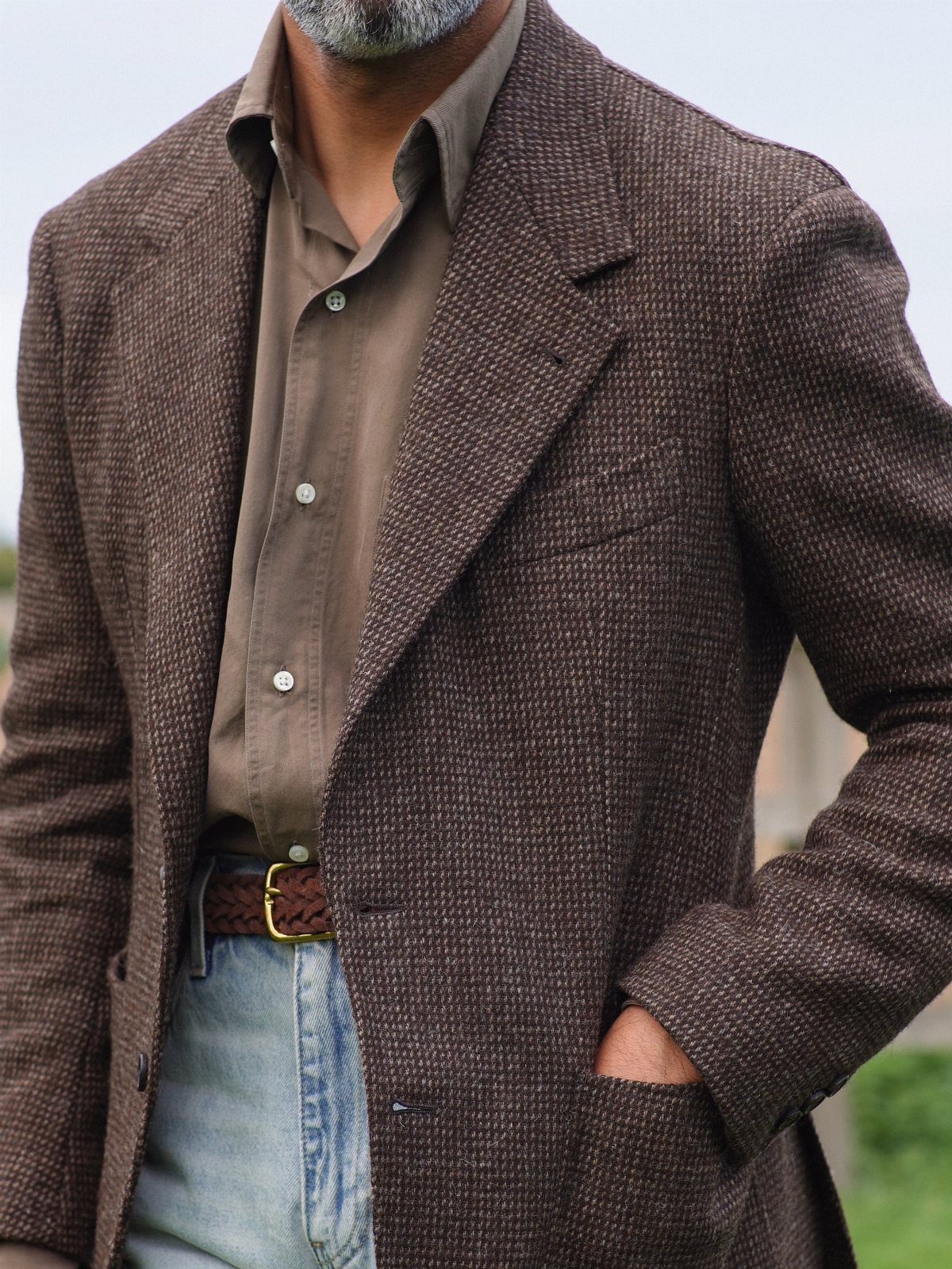 Man wearing a brown textured blazer, brown shirt, and light blue denim jeans with a braided belt.