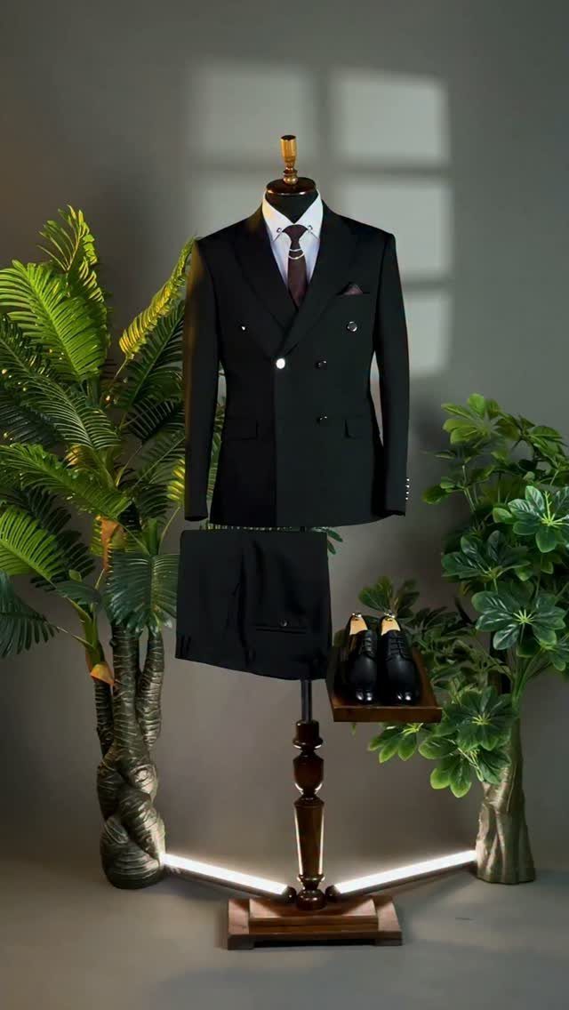 A black double-breasted suit and dress shoes displayed on a mannequin stand with green plants.