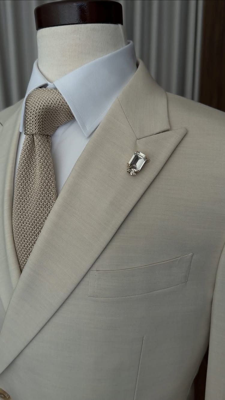 A beige suit jacket with a textured tan tie and a silver lapel pin on a mannequin.