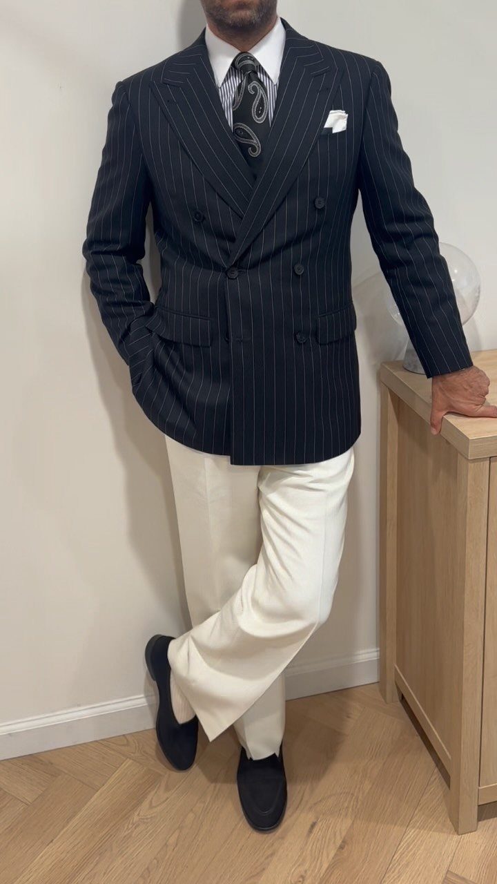 A man in a navy pinstripe blazer, white shirt, patterned tie, and white trousers.
