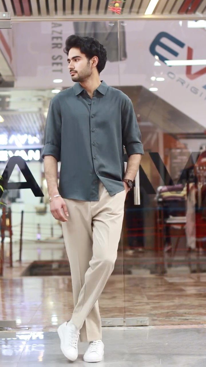 A man wearing a teal button-down shirt, beige trousers, and white sneakers standing outdoors.