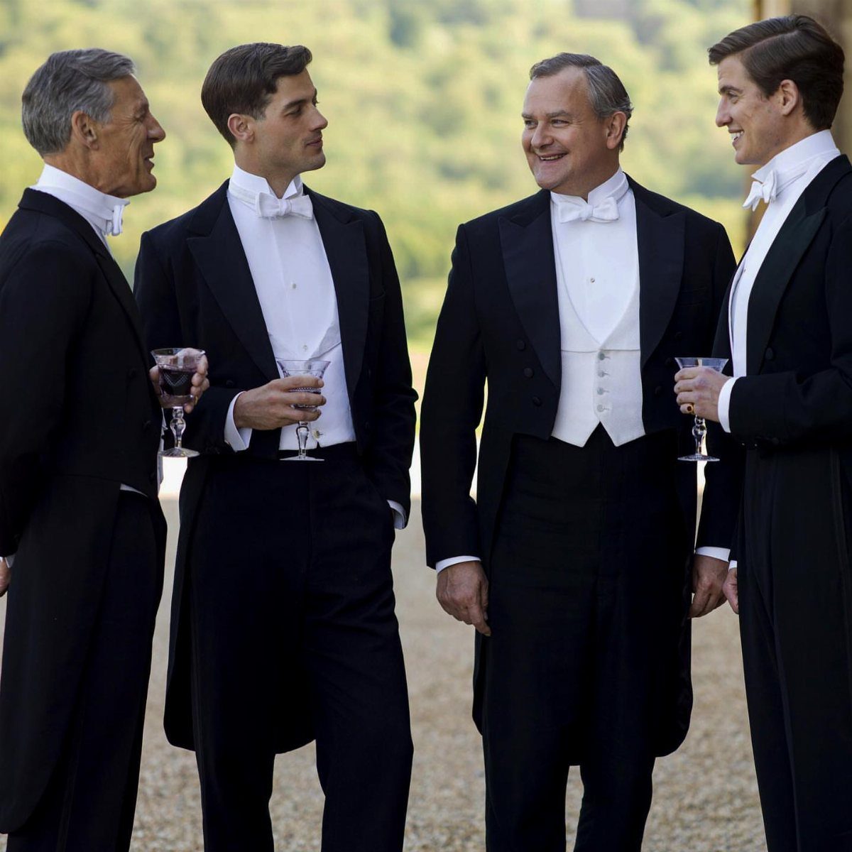 Four men dressed in formal white tie attire standing together outdoors holding drinks.