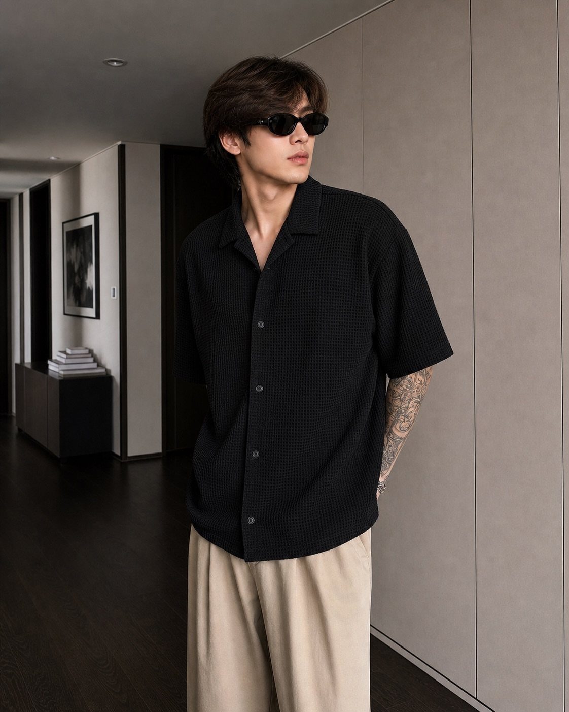 A young man wearing a black short-sleeved button-up shirt and sunglasses in a hallway.