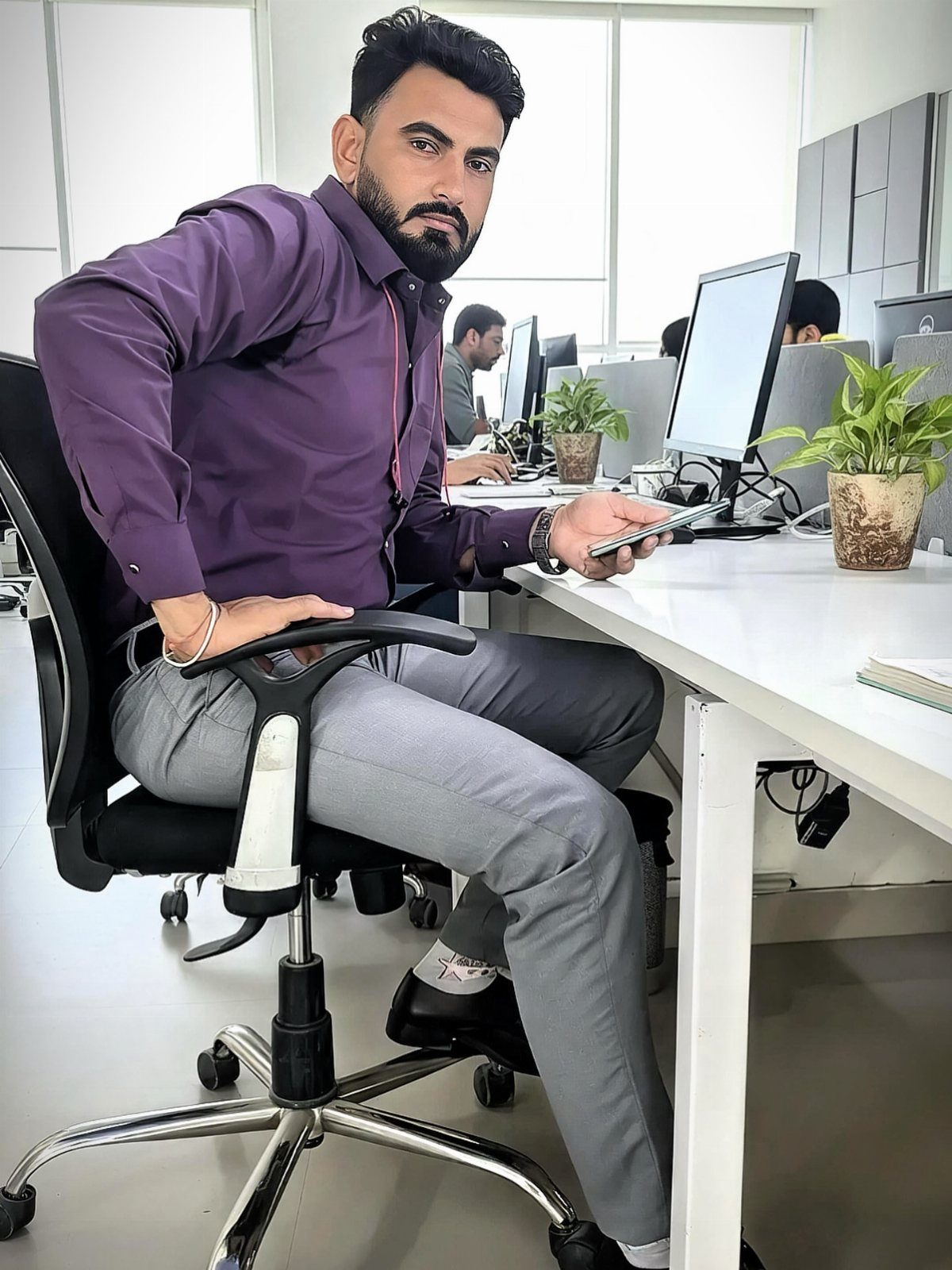 A man in a purple shirt and grey trousers sitting at a white office desk.