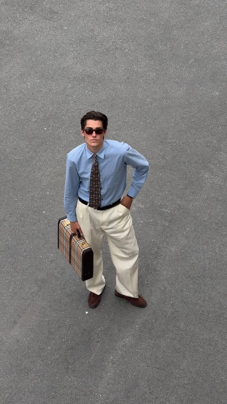 Man in light blue shirt, patterned tie, cream trousers, and sunglasses holding a vintage briefcase.