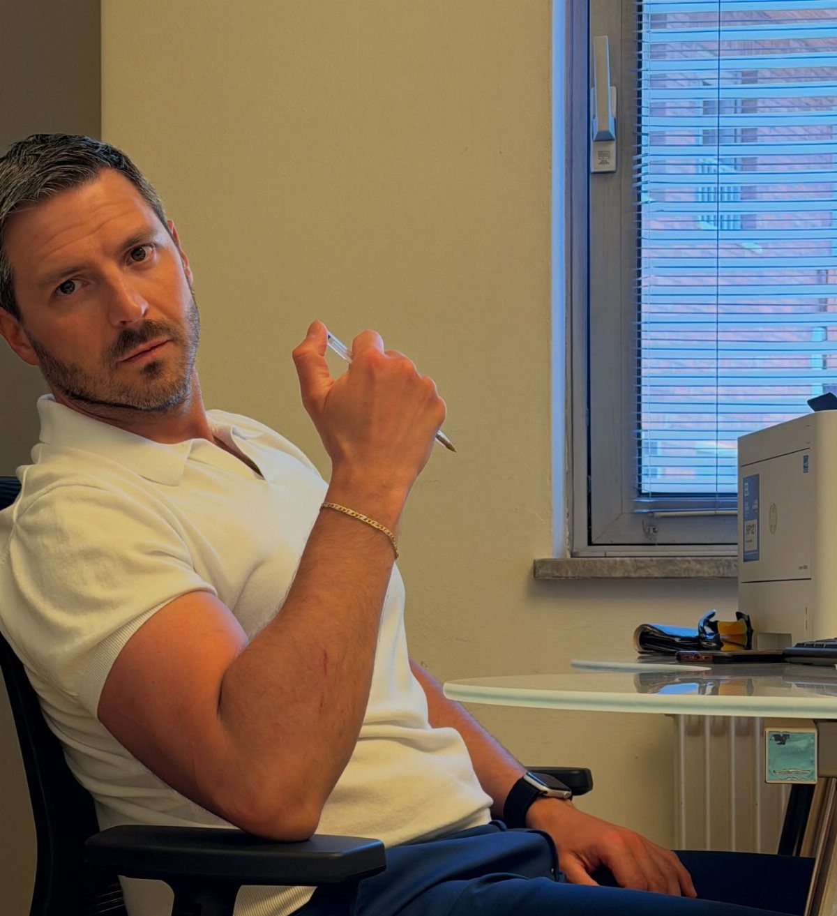 A man in a white polo shirt sitting in an office chair holding a pen.