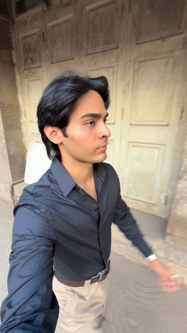 A young man in a black button-down shirt and beige trousers standing by a wooden door.