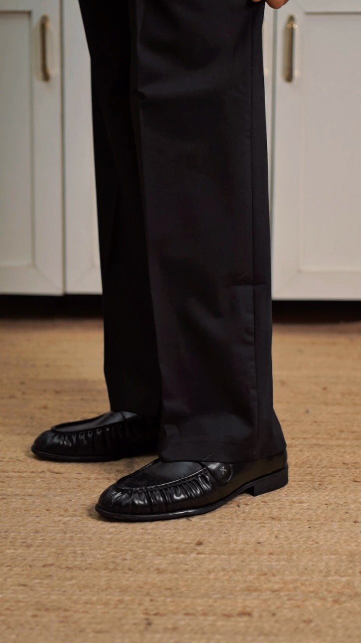Black leather loafers worn with black trousers on a tan carpeted floor.