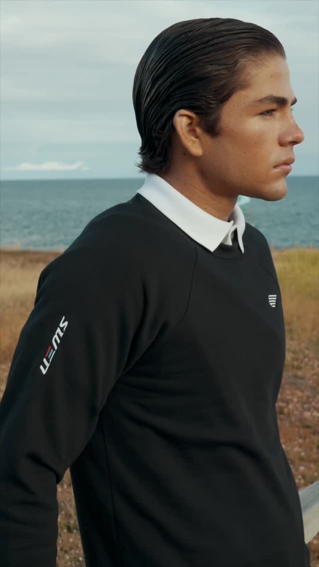 A young man wearing a black sweatshirt over a white collared shirt by the sea.