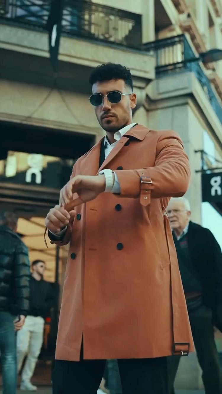 A man in a tan trench coat and sunglasses looking at his watch on a city street.