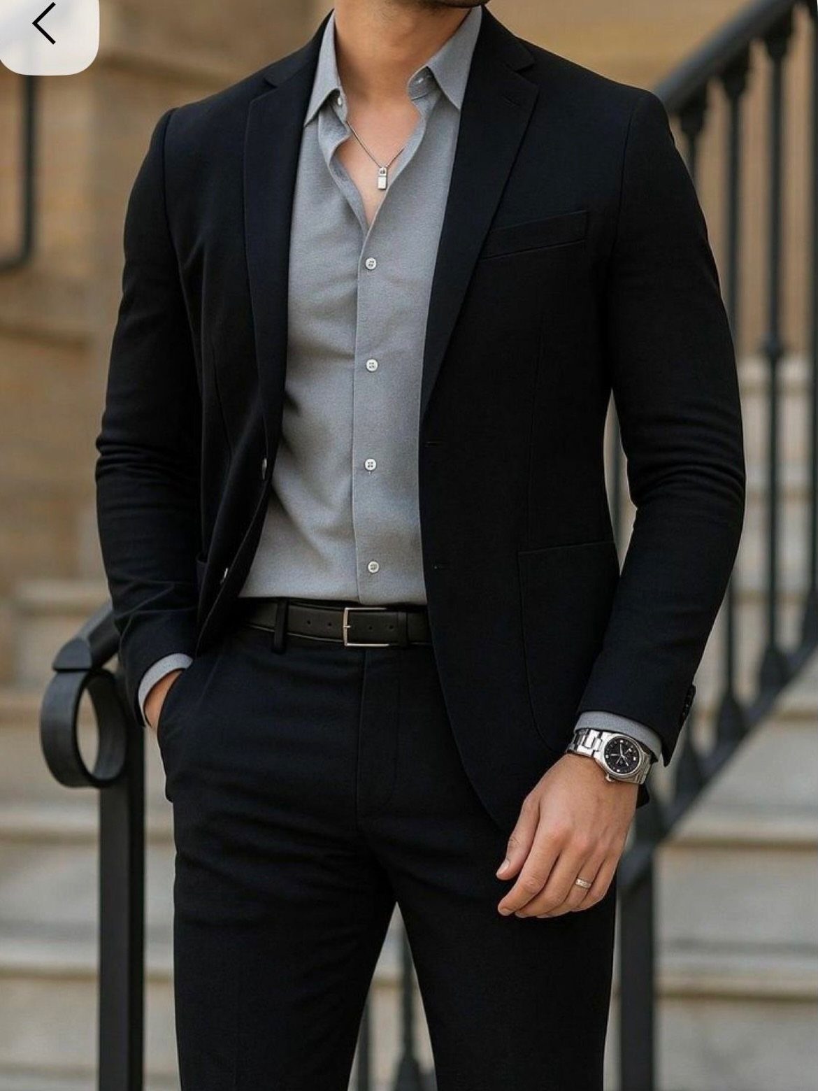Man wearing a black blazer, grey button-down shirt, and black trousers standing outdoors.