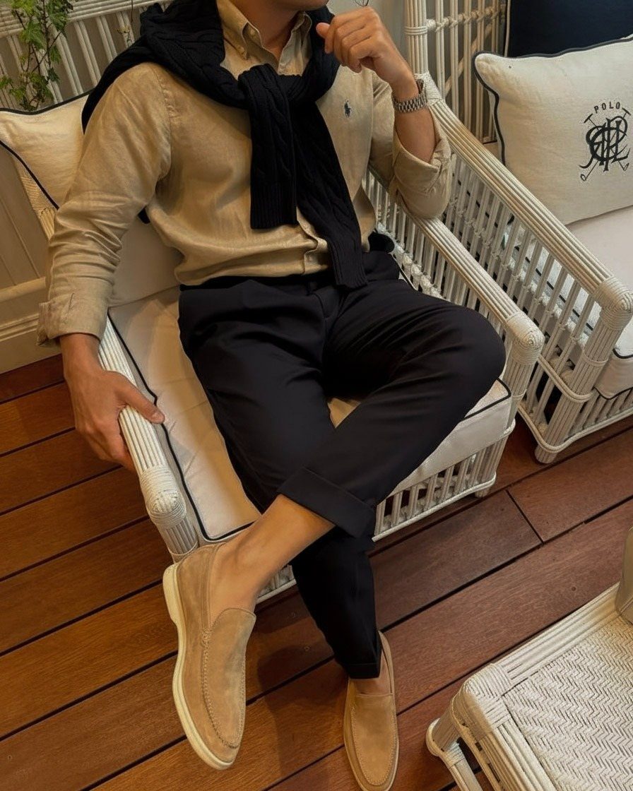 Man in tan shirt and black trousers wearing tan suede slip-on shoes sitting outdoors.